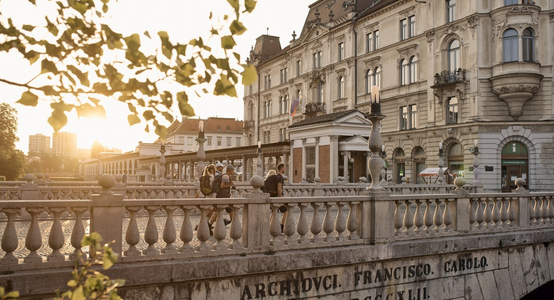 A cityscape with a historical building and a group of people walking along a promenade during sunset, with trees in the foreground.