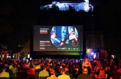 Gledalci sedijo na stolih, postavljenih na trgu v srediscu mesta, in gledajo film na velikem platnu na prostem.
