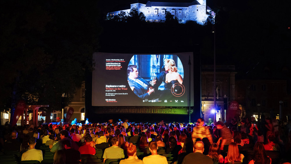Viewers are seated on chairs set up in the town square, watching a film on a large outdoor screen.