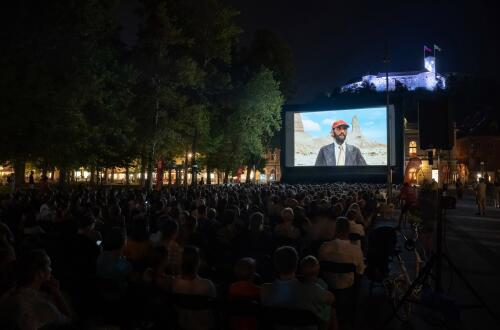 Obcinstvo spremlja projekcijo filma na prostem z osvetljenim Ljubljanskim gradom v ozadju.
