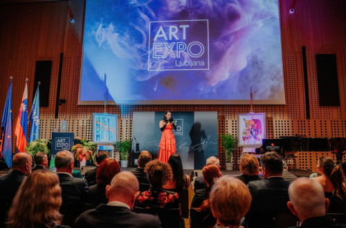 Na odru stoji zenska v rdeci obleki, pred njo publika, ona pa povezuje dogodek.