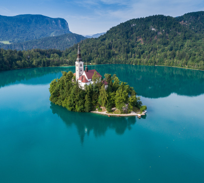 Majhen otok s cerkvijo sredi smaragdno zelenega Blejskega jezera, obdan z gozdnatimi hribi.