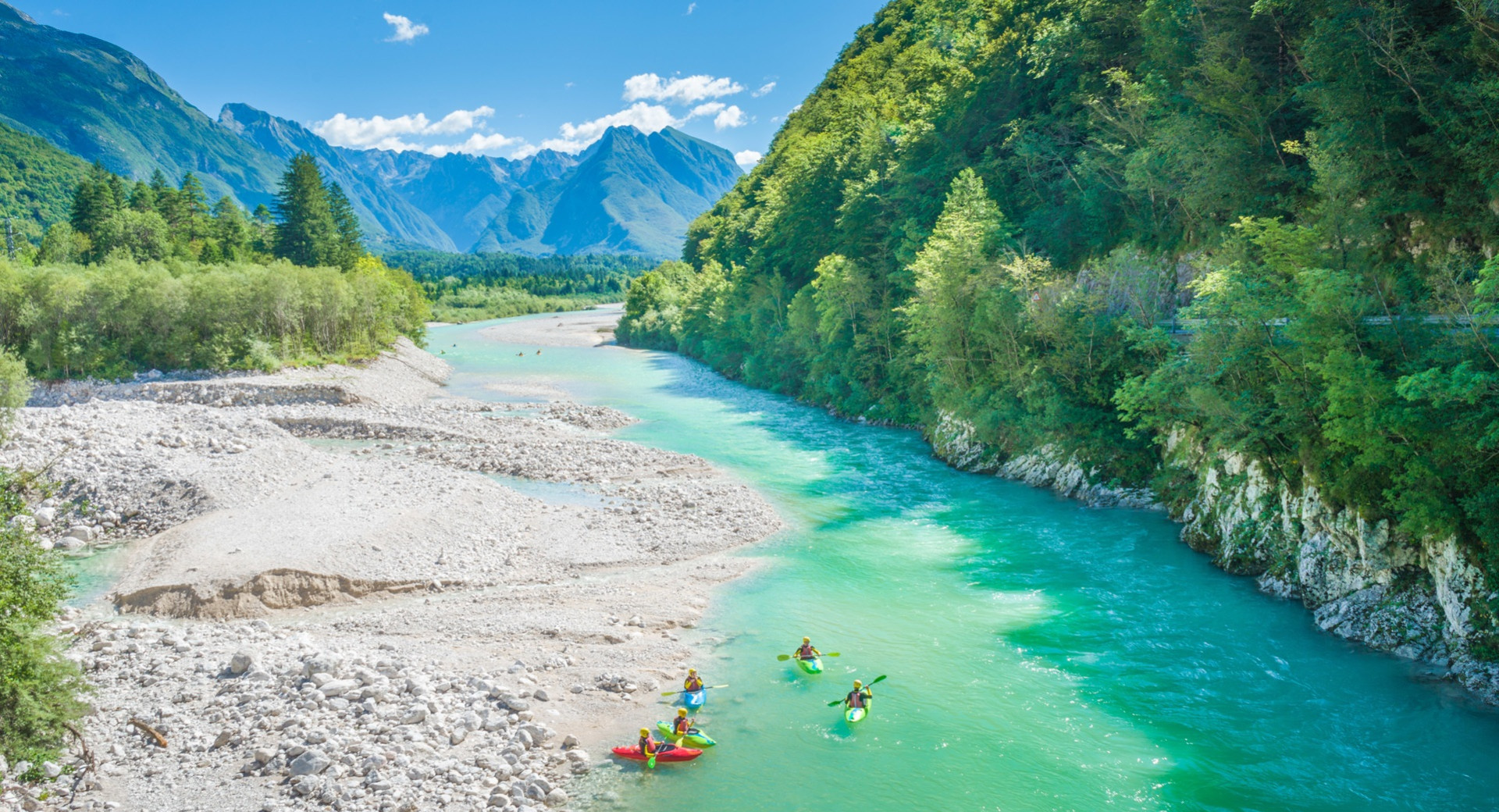 A group of kayakers enjoys paddling along an emerald-green river, surrounded by majestic mountains and dense forest.
