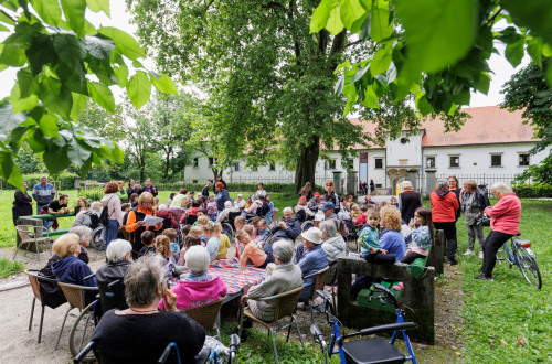 Children and older people mingle at an intergenerational event in a park by a white building with a red roof.
