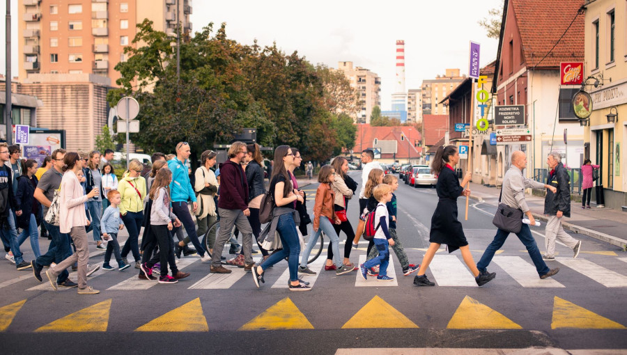 A group of people of different ages crosses a city pedestrian crossing in an urban setting, surrounded by residential and commercial buildings.