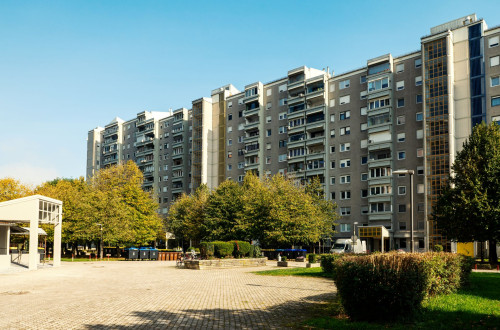Stanovanjski blok z vec nadstropji in balkoni, obdanim z drevesi, grmovjem in urejenim dvoriscem na soncen dan.