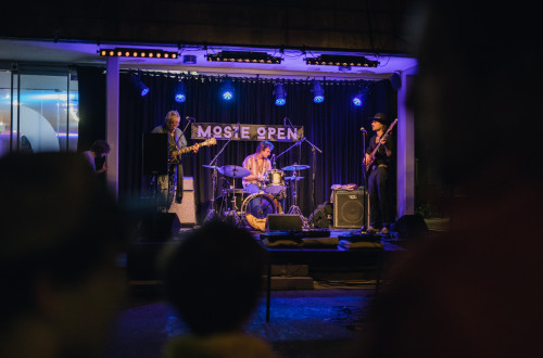An evening music group performing live on stage at the Moste Open event, accompanied by lights and a foreground audience.