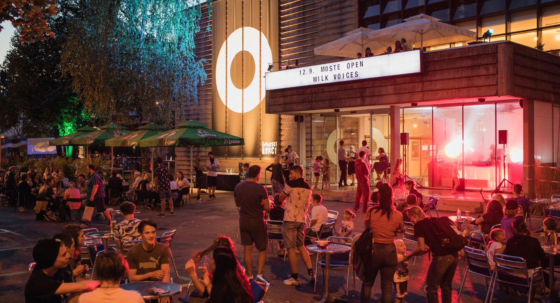 An evening street event, Moste Open, taking place in front of a cultural centre with a DJ, lighting, and outdoor socialising.