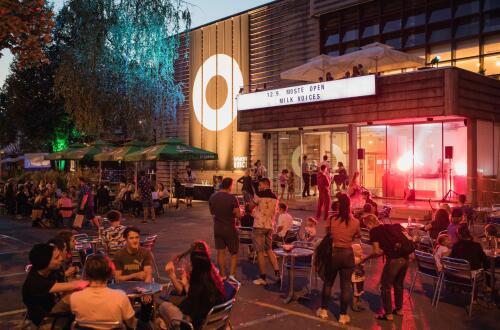 Vecerni ulicni dogodek Moste Open pred kulturnim centrom z DJ-jem, osvetlitvijo in druzenjem obiskovalcev na prostem.