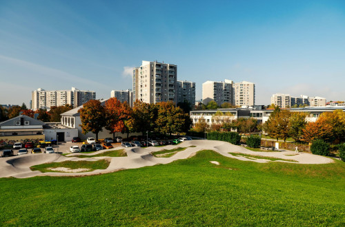Zelen park s pumptrack stezo, parkiranimi avtomobili in vecstanovanjskimi bloki v ozadju pod jasnim modrim nebom.