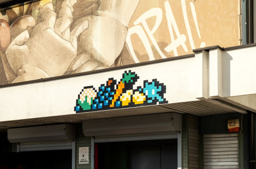 Mozaicna ulicna umetnost s pikseliziranim motivom sadja in zelenjave na procelju stavbe s stensko poslikavo in napisom »OPA!«.
