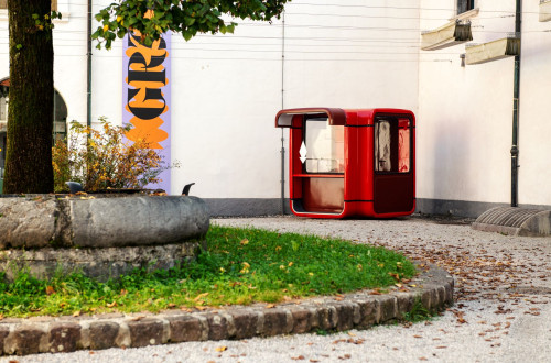 Rdec ulicni kiosk ob kamnitem vodnjaku na dvoriscu z zelenjem in zastavo kulturnega dogodka v ozadju.