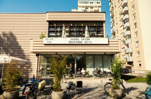 Ljudje sedijo pred kulturnim centrom z napisom o dogodku Zavod Sploh (Ne)forma #113, obdanem z bloki in kolesi.