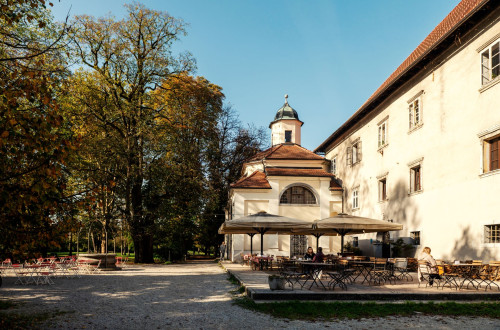 A sunny terrace with vintage tables next to a historic building and a chapel, surrounded by autumn trees and a peaceful park atmosphere.