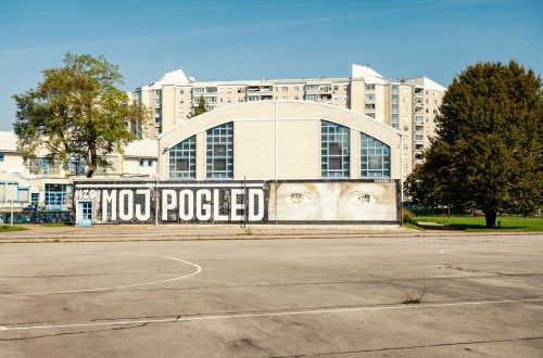 A mural with the words “Moj pogled” and eyes on the façade of a sports hall next to a playground in a residential neighbourhood.