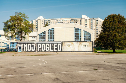 Stenska poslikava z napisom »Moj pogled« in ocmi na fasadi sportne dvorane ob igriscu v stanovanjskem naselju.
