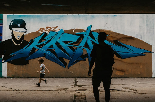 Grafit na steni podhoda; mimo tece decek, v senci stoji moski.