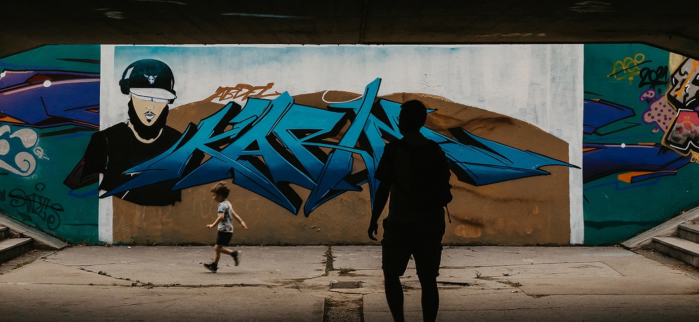 Graffiti on the underpass wall; a boy runs past, a man stands in the shadows.