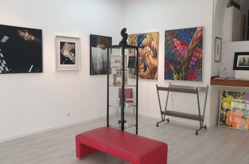 Svetla galerija z barvitimi slikami na belih stenah, stekleno vitrino na sredini prostora in rdeco klopjo v ospredju.