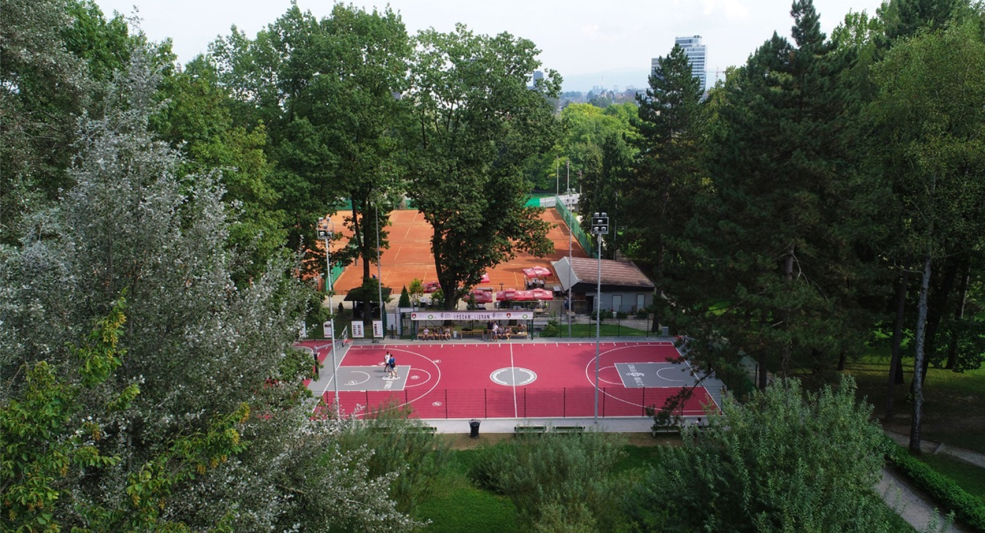 A sports court with a red surface surrounded by greenery.