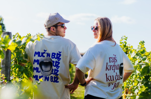 A man and a woman wearing graphic slogan T-shirts stand among sunlit vineyard rows, looking back over their shoulders at each other.