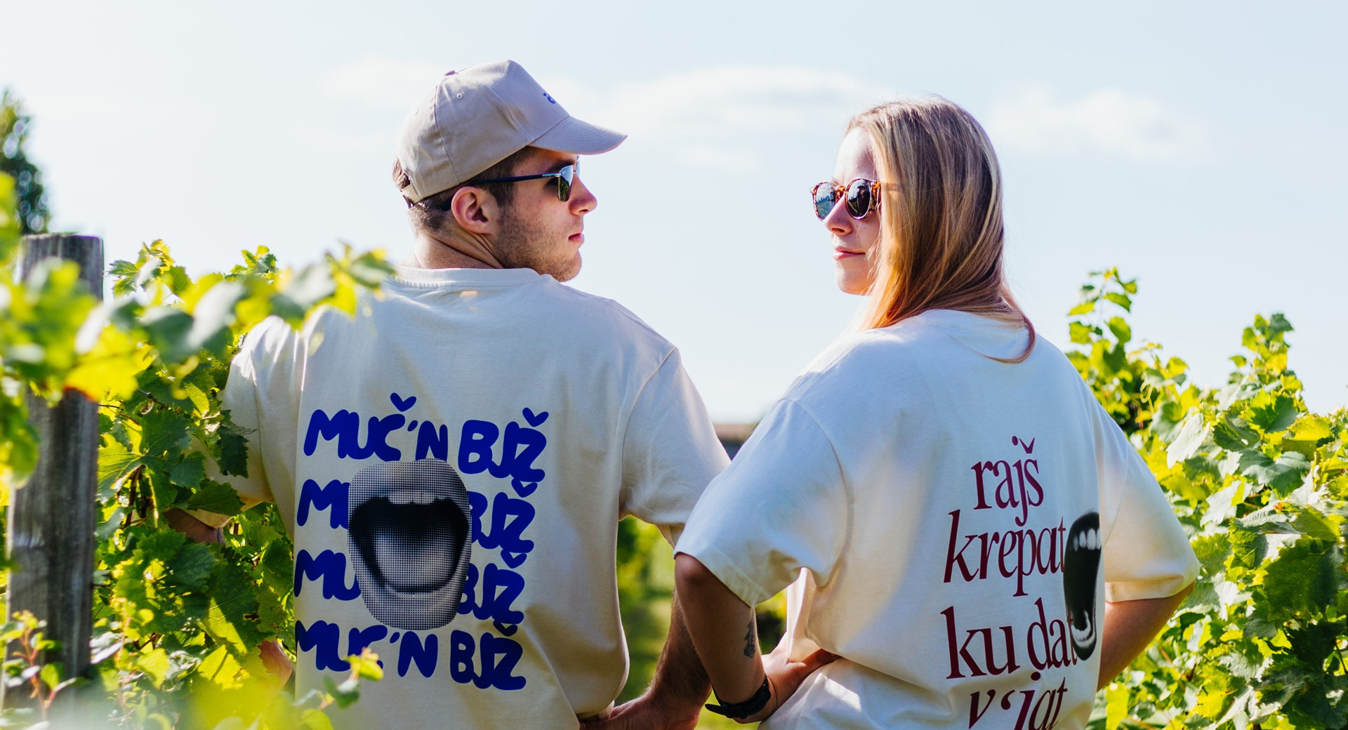 Moski in zenska v majicah z graficnimi slogani stojita med soncnimi vinogradniskimi vrstami in se ozreta nazaj drug proti drugemu.