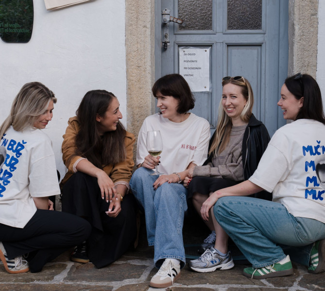 Pet zensk sedi skupaj na pragu, se smeji in pogovarja, ena izmed njih drzi kozarec belega vina.