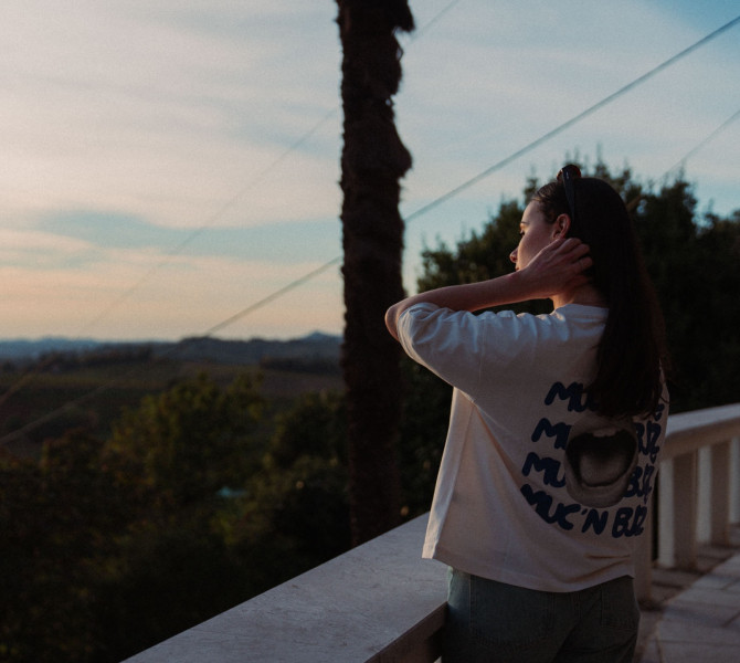 Zenska v majici z graficnim sloganom stoji na terasi ob soncnem zahodu in gleda cez slikovito vinogradnisko pokrajino.