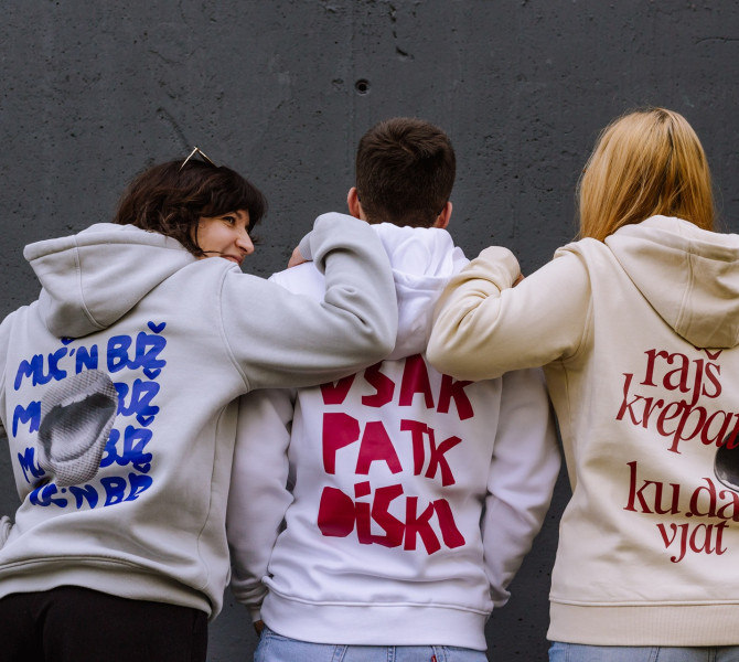 Trije ljudje stojijo s hrbtom proti kameri, nosijo puloverje s kapuco z izrazitimi graficnimi potiski in besedilom ter se drzijo cez ramena pred preprosto steno.