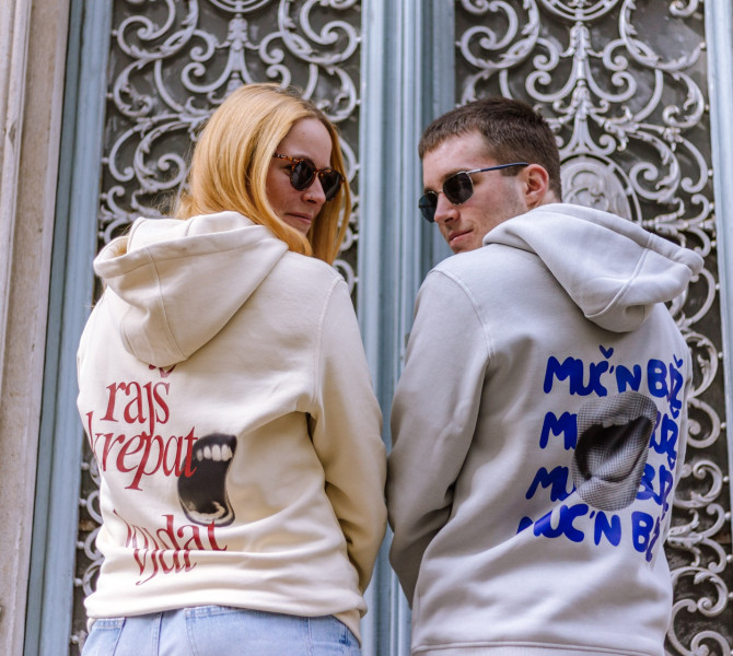 Dve osebi s soncnimi ocali gledata nazaj cez ramo, nosita puloverje s kapuco z velikimi graficnimi napisi in stojita pred okrasenimi kovinskimi vrati.