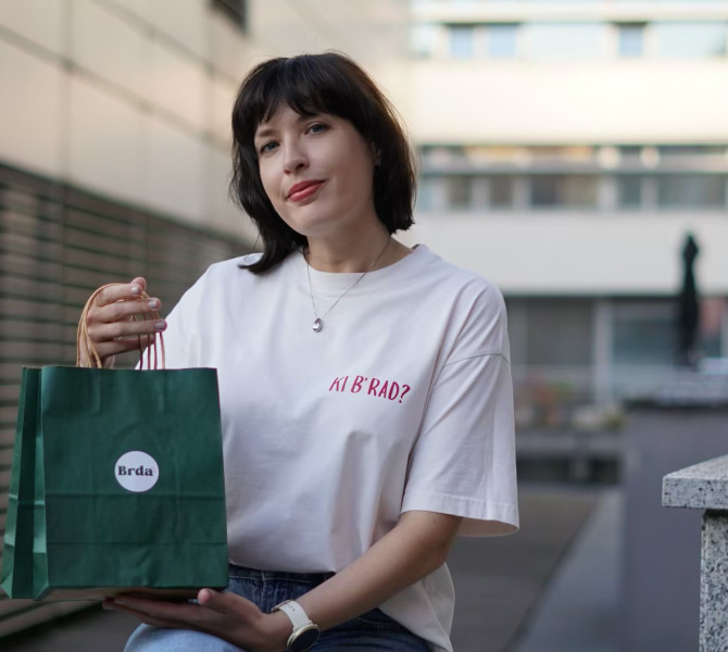 Zenska v minimalistcni beli majici z igrivim sloganom sedi in drzi zeleno nakupovalno vrecko z napisom Brda v sodobnem urbanem okolju.
