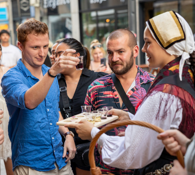 A man in a blue shirt has taken a glass of drink from a wooden tray held by a woman in traditional costume and is holding it in front of him, looking at it, while other people are around them.