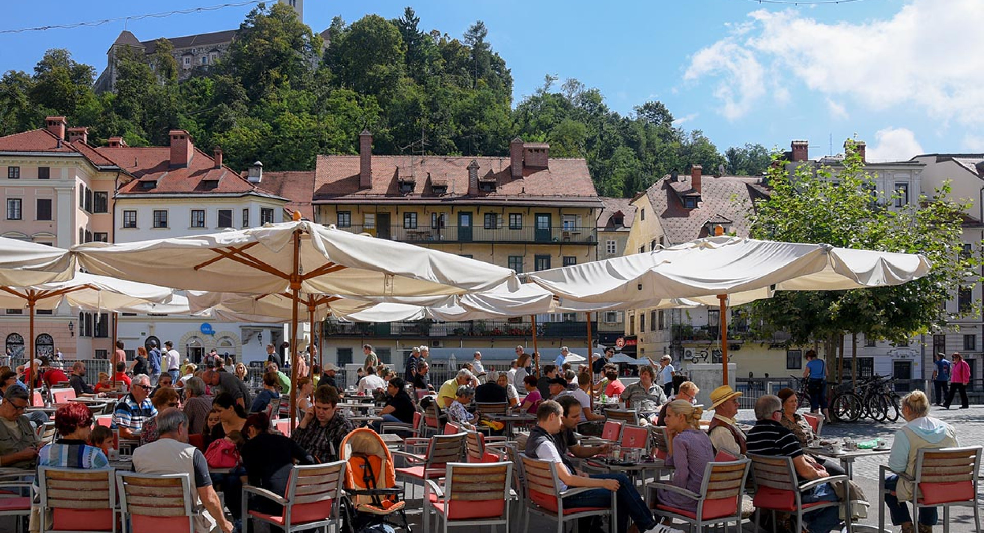 Obiskovalci na zunanji terasi pod senčniki. V ozadju Ljubljanski grad.