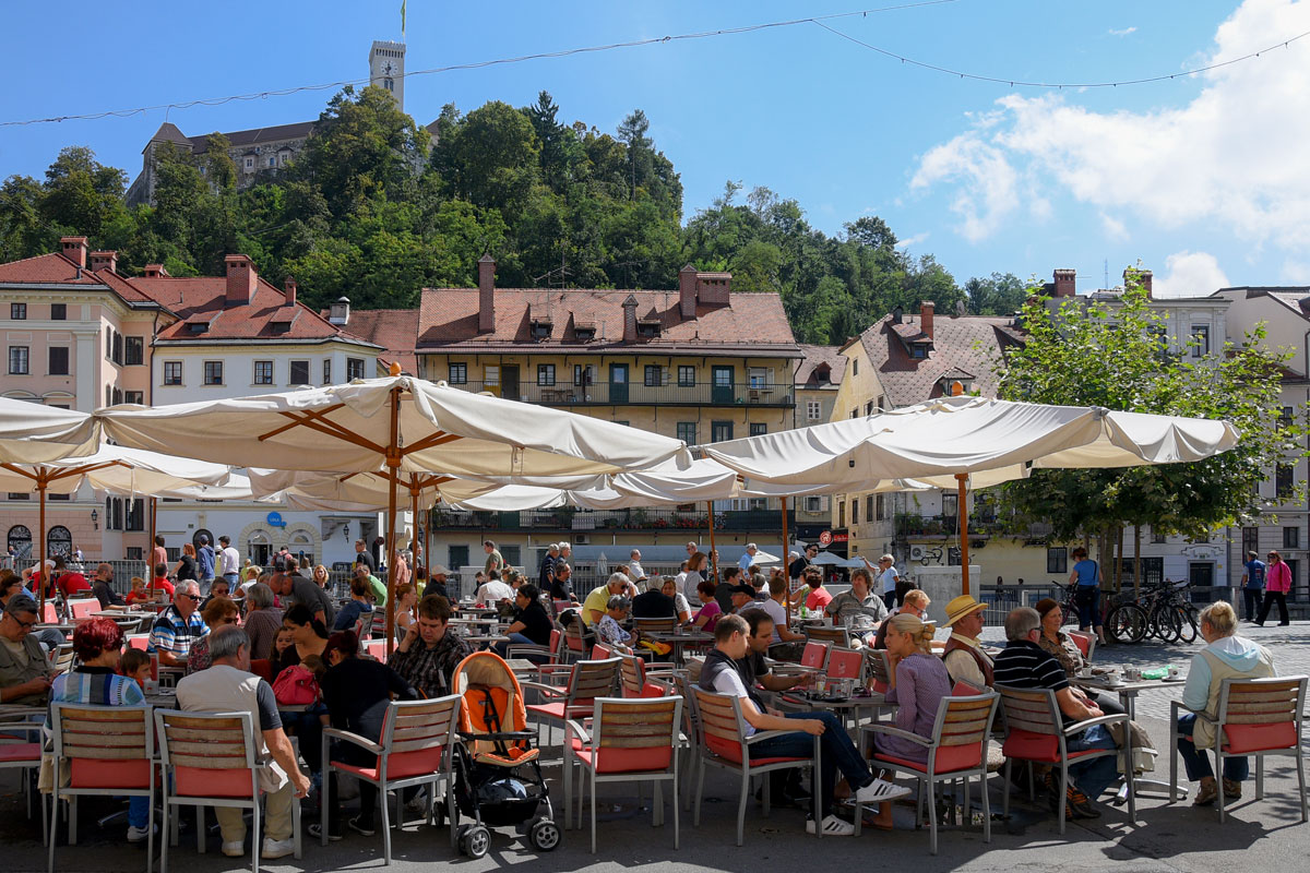 Obiskovalci na zunanji terasi pod senčniki. V ozadju Ljubljanski grad.