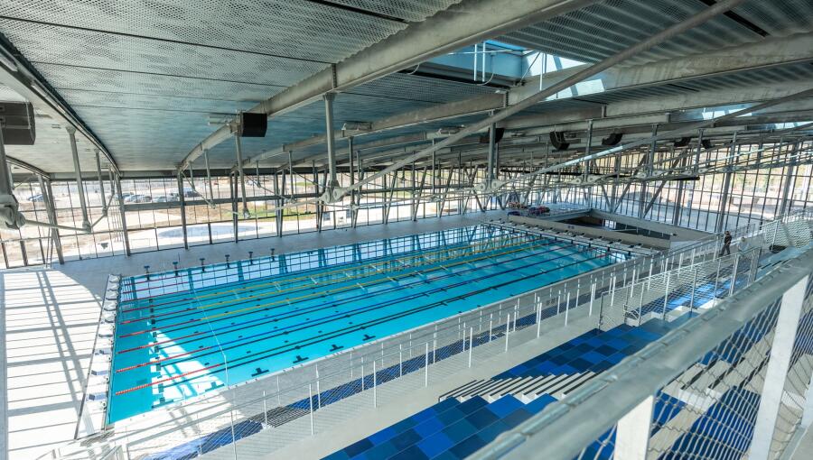 The image shows a spacious indoor swimming pool with bright blue water and marked lanes. It has a modern structure with large windows allowing natural light to enter, creating a vibrant atmosphere.