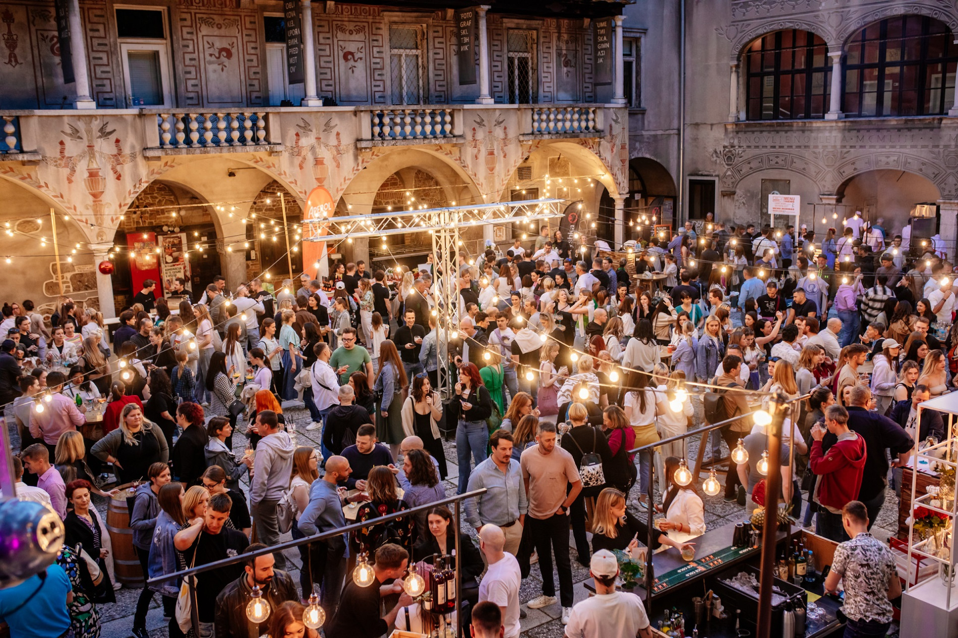 Skupina ljudi se v mraku druzi v ljubljanskih Krizankah, v domacem ambientu ob toplih rumenih luceh.