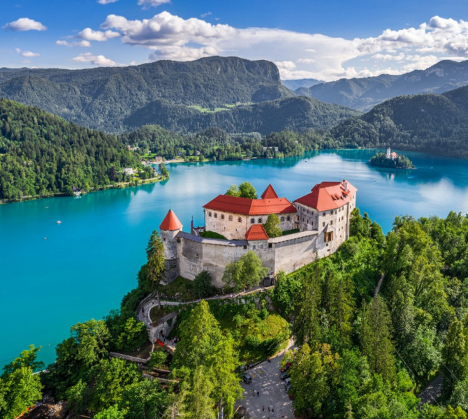 Bled Castle perched on a forested hill overlooking the turquoise waters of Lake Bled, with the island and surrounding mountains in the distance.