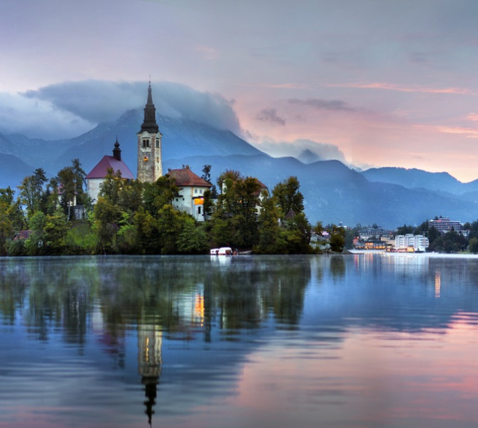 Island church with tall steeple on calm lake at dawn, mountains in background, all reflected on the water.
