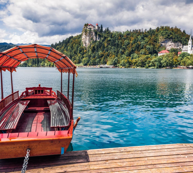 Traditional wooden boat moored at the dock on Lake Bled, with Bled Castle and a church visible on the forested hills in the background.
