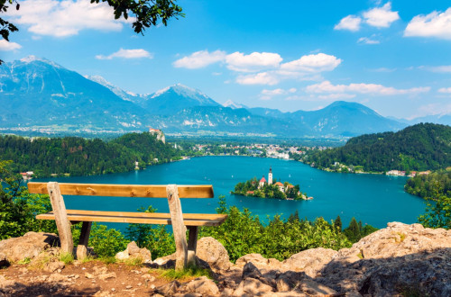 Wooden bench on a hill overlooking Lake Bled with the island church and Alpine mountains in the background.