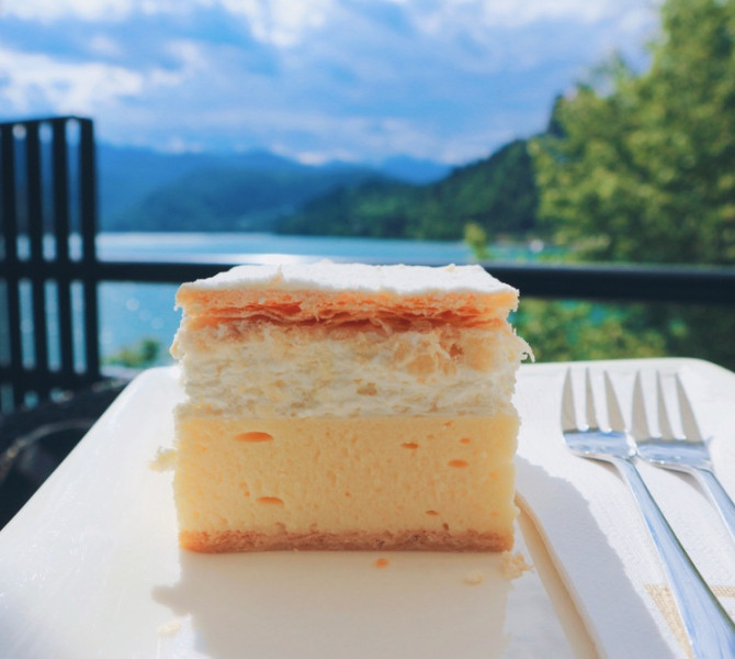 Slice of cream cake on a plate with fork, overlooking a lake and forested hills in the background.
