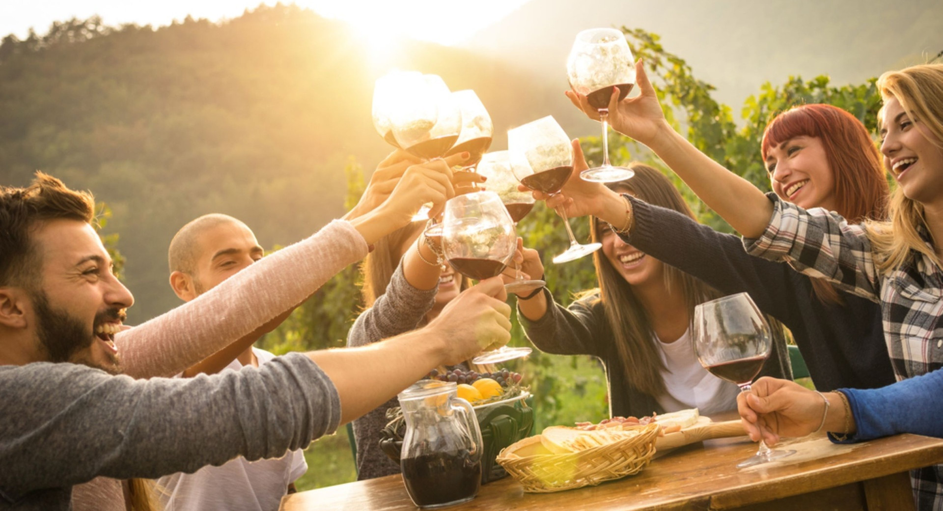 Friends toasting with glasses of red wine at a vineyard table in the warm glow of sunset.