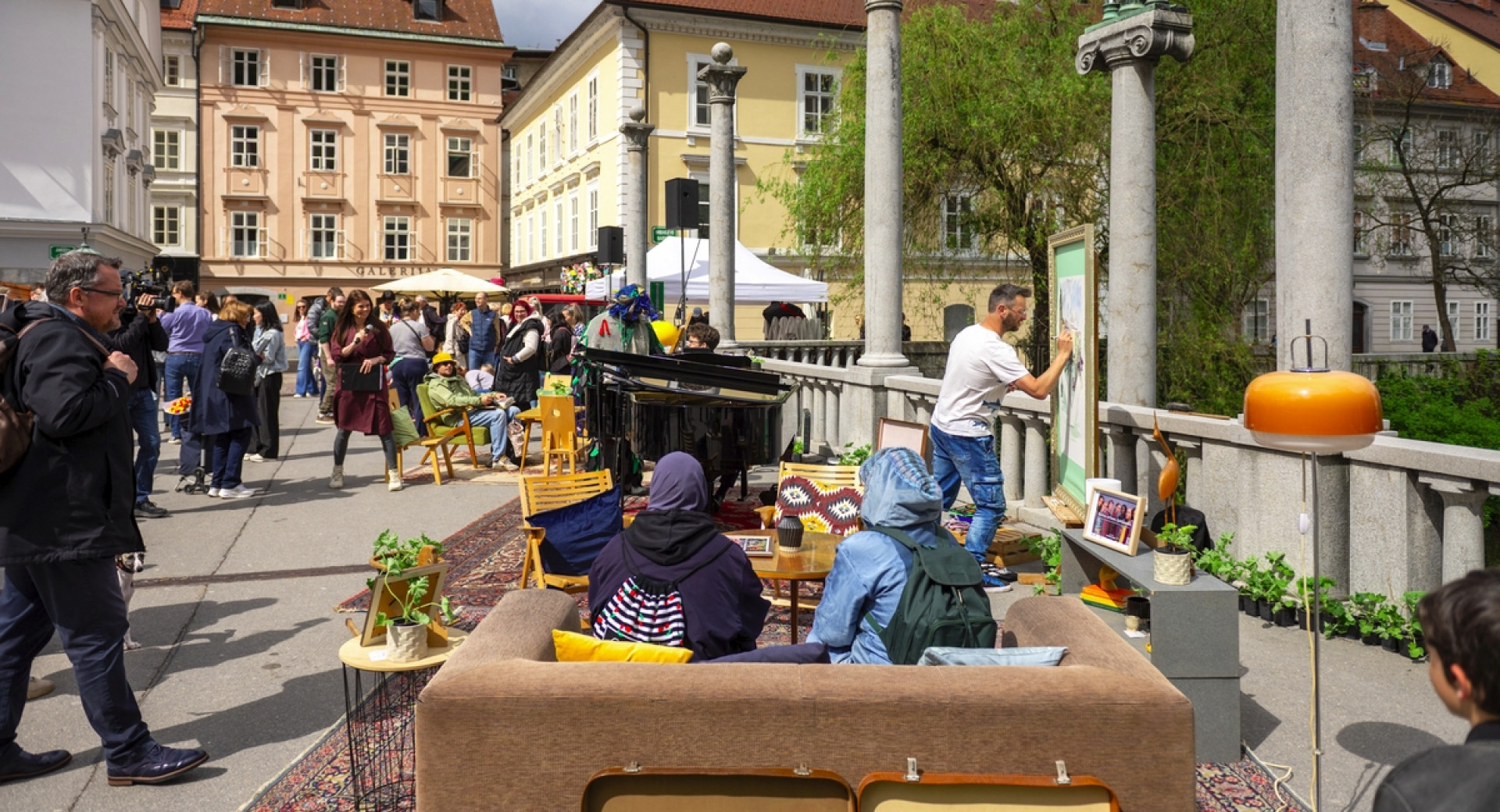 Dogajanje na prostem: obiskovalci sedijo na kavču, umetnik riše na platno, pianist igra na klavir ...