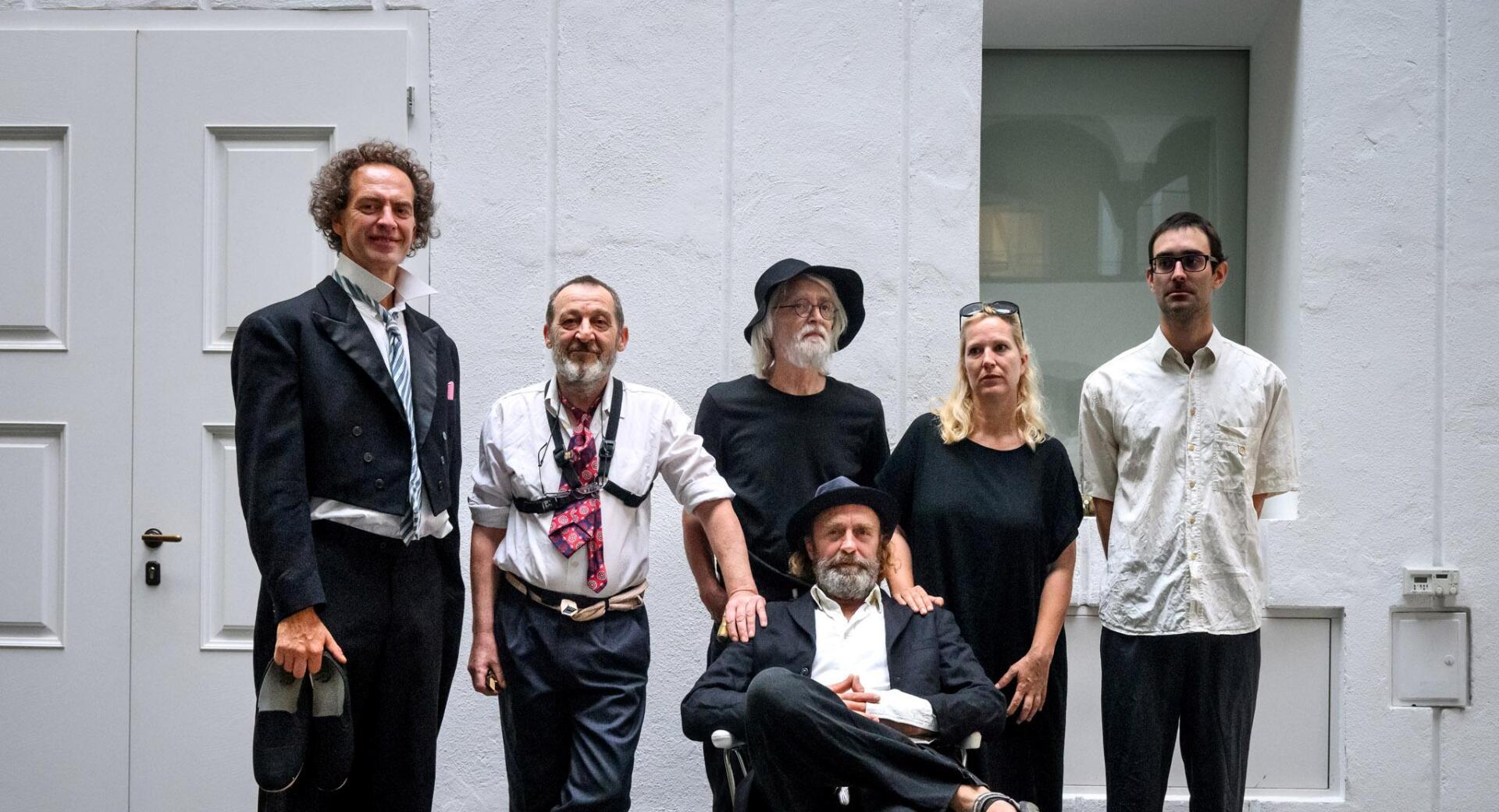 A group of six people poses in a minimalist white interior space, with one man seated in a chair in front while the others stand around him in casual, artistic portrait composition.