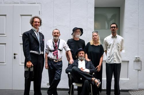 A group of six people poses in a minimalist white interior space, with one man seated in a chair in front while the others stand around him in casual, artistic portrait composition.