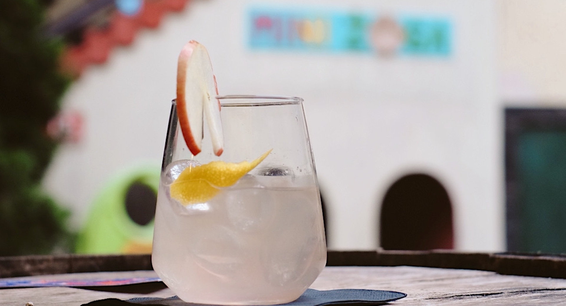 A glass of cloudy liquid adorned with slices of apple and lemon. The glass is on a wooden tray, with a building and bushes out of focus in the background.