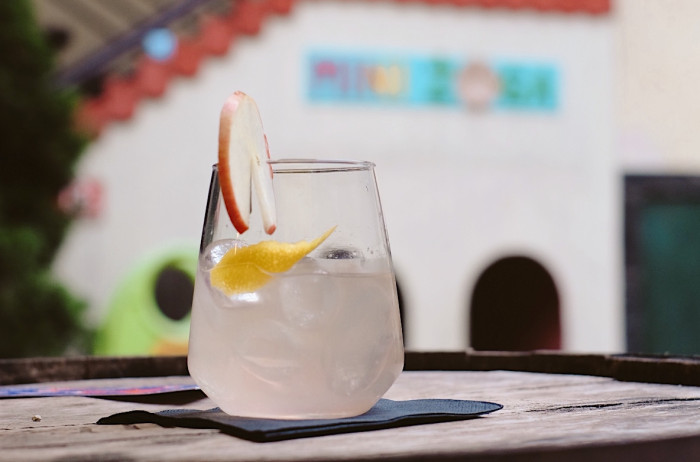 A glass of cloudy liquid adorned with slices of apple and lemon. The glass is on a wooden tray, with a building and bushes out of focus in the background.