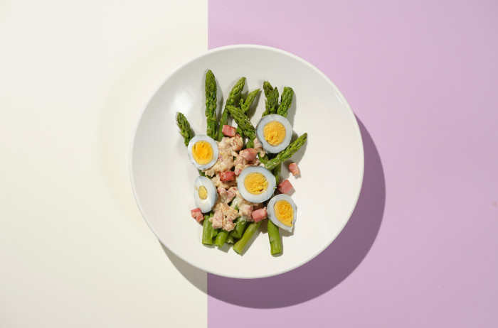 A white plate on a beige and violet background. The plate features neatly presented asparagus with chopped hard-boiled eggs, sprinkled with prosciutto.