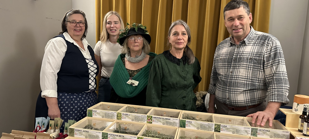 Skupina petih ljudi stoji za mizo na sejmu. Na mizi so razstavljene številne lesene škatle s posušenimi zelišči in čaji.