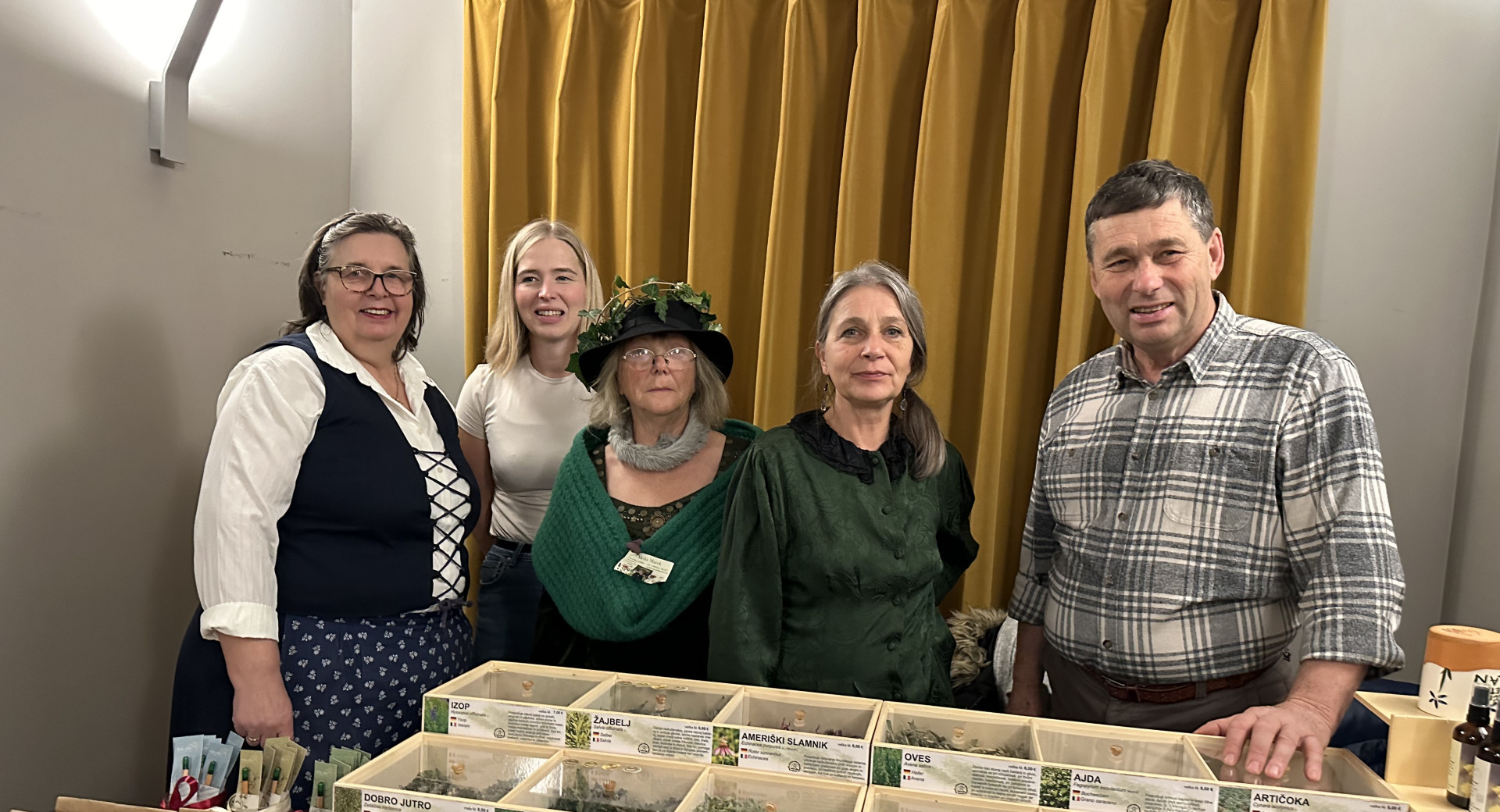 A group of five people is standing behind a table at a fair. Numerous wooden boxes with dried herbs and teas are displayed on the table.